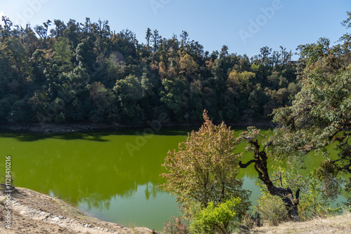 A serene green lake is surrounded by a dense forest of lush trees in varying shades of green and autumn hues. The vibrant foliage contrasts beautifully with the calm, reflective waters of deoria lake. photo