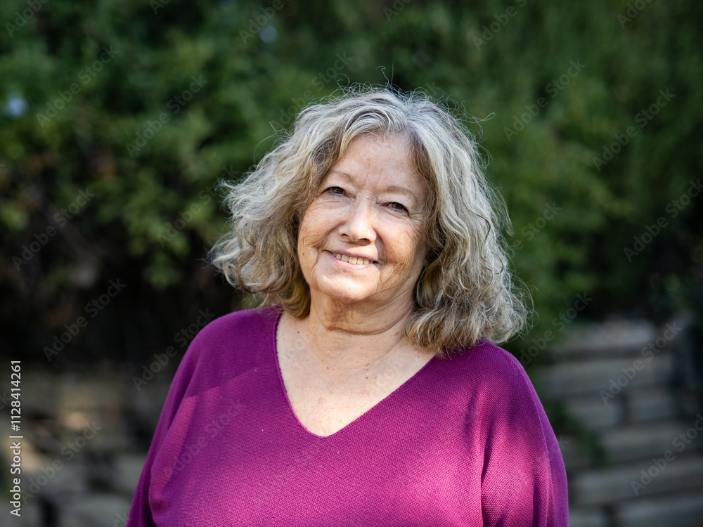 A cheerful elderly woman stands outdoors, wearing a purple sweater. Her curly gray hair frames her face as she smiles warmly, surrounded by lush greenery. A cheerful elderly woman stands outdoors, wearing a purple sweater. Her curly gray hair frames her face as she smiles warmly, surrounded by lush greenery.