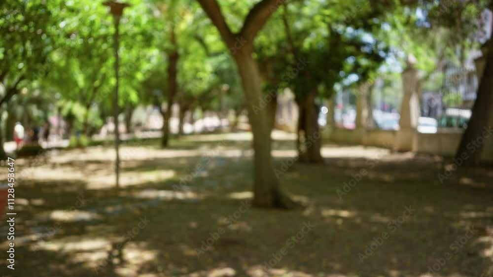 Defocused outdoor park scene with blurred trees and bokeh background, featuring out of focus individuals walking under shaded pathways in a lush green setting on a sunny day Defocused outdoor park scene with blurred trees and bokeh background, featuring out of focus individuals walking under shaded pathways in a lush green setting on a sunny day