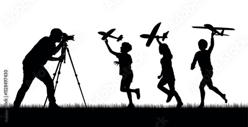 Man with camera shooting videos of children running while holding airplane toy outdoor on grass field silhouettes.
