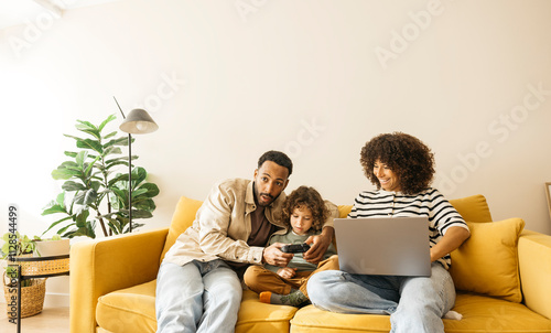 Foto Family members relaxing on a bright yellow sofa and playing video games while the mother is using a laptop