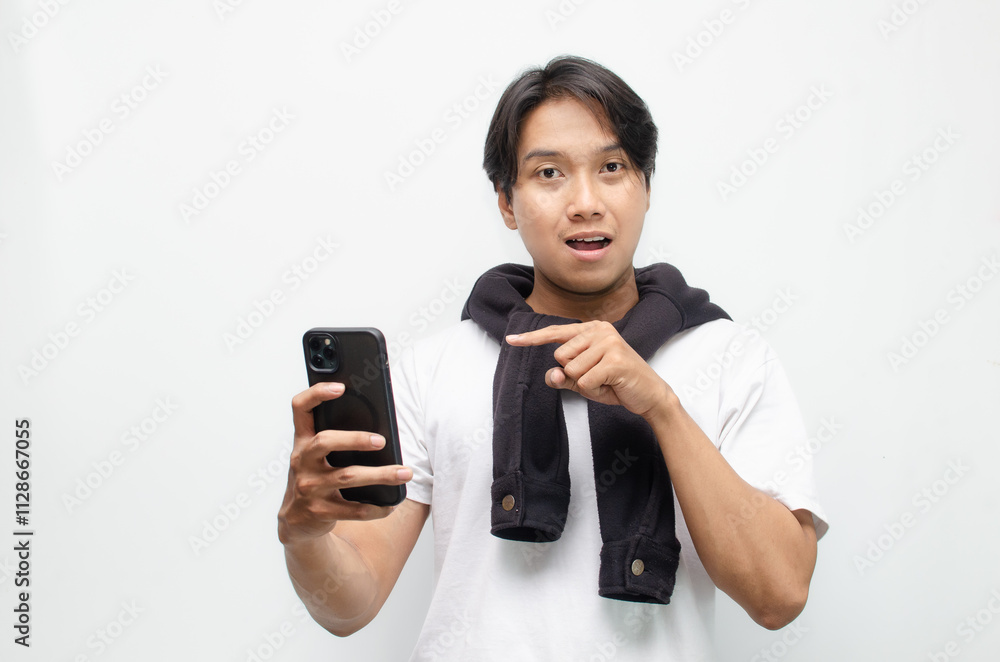 Excited Young Asian Man in Casual Outfit Holding Smartphone and Pointing with Joyful Expression Excited Young Asian Man in Casual Outfit Holding Smartphone and Pointing with Joyful Expression