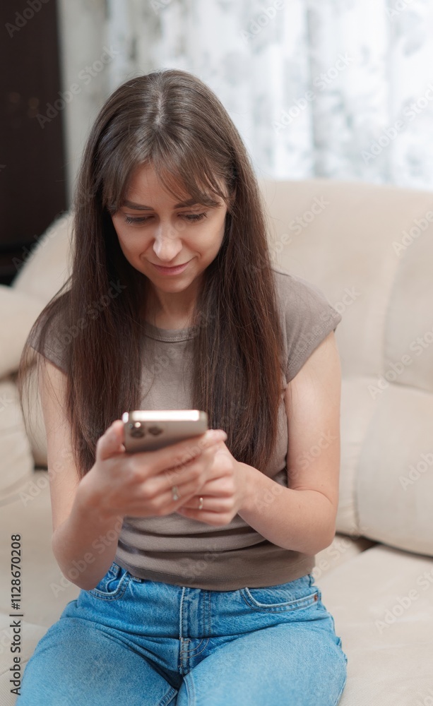 Young happy woman sitting comfortably on sofa, smiling while using her phone indoors during a relaxing afternoon vertical