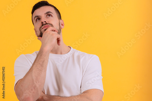 Canvastavla A young man is standing against a bright yellow background, looking up thoughtfully with a hand on his chin