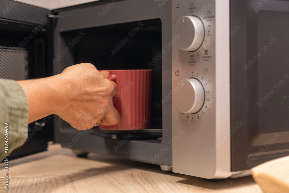 Fototapeta premium Female hand places a red mug inside a microwave