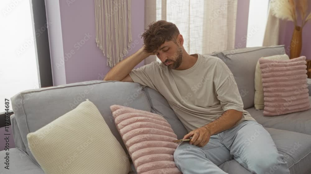 Young man sitting on a couch looking at his phone with a sad expression in a stylishly decorated living room. Young man sitting on a couch looking at his phone with a sad expression in a stylishly decorated living room.