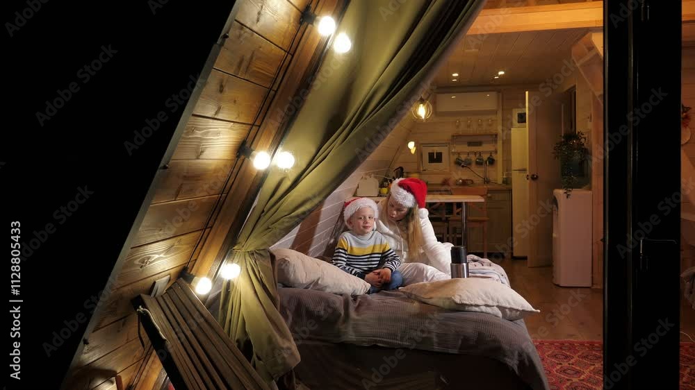 A triangular wooden house with a large window and small lights in the evening, a mother and son in Christmas hats drinking tea on a cold winter evening. A triangular wooden house with a large window and small lights in the evening, a mother and son in Christmas hats drinking tea on a cold winter evening.