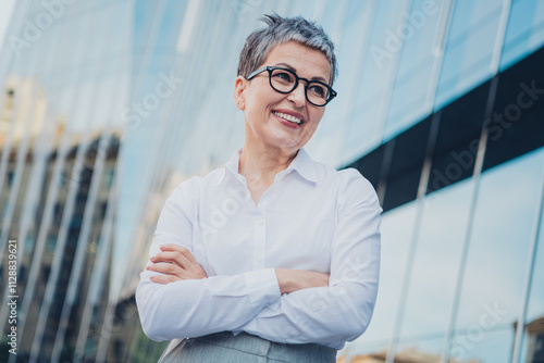 Confident middle-aged businesswoman with short grey hair and glasses outdoors in urban cityscape, wearing elegant white shirt.