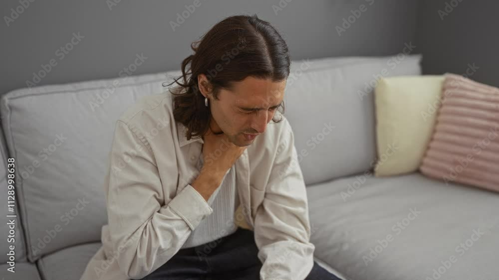 Young man with long hair holding his throat and coughing in a living room, indicating a sore throat at home Young man with long hair holding his throat and coughing in a living room, indicating a sore throat at home