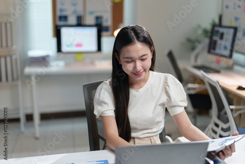 Young office worker analyzing marketing data and financial reports holding folder with documents and using laptop computer