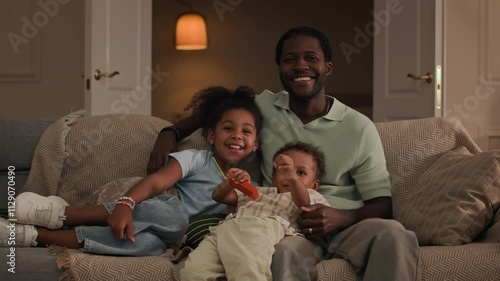 Medium shot of caring young African American father, his toddler son and little daughter sitting together on couch, hugging each other and enjoying watching TV in cozy modern living room