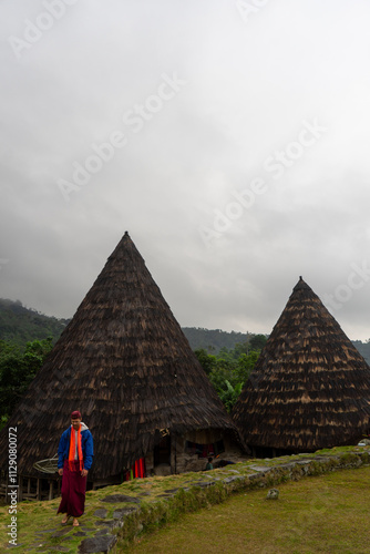 A person stands near traditional cone-shaped houses in a lush, misty landscape. photo