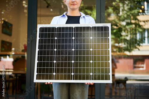 Woman holding solar panel for charging, stands outdoors in urban setting. Cropped view. Concept of green energy generation. photo