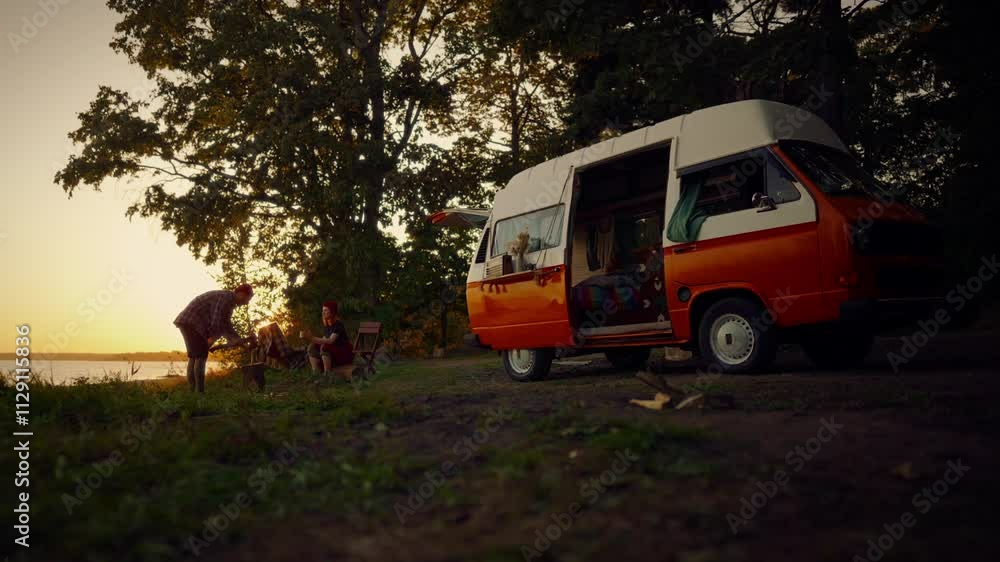Pair of tourist spending summer vacation in nature, traveling by camper van. Man and woman chopping wood for campfire, halt on shore of beautiful river in evening, picturesque landscape in sunset Pair of tourist spending summer vacation in nature, traveling by camper van. Man and woman chopping wood for campfire, halt on shore of beautiful river in evening, picturesque landscape in sunset