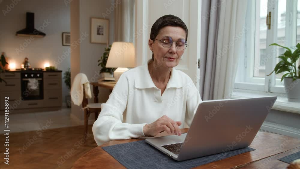 Senior elegant woman sits at table typing on keyboard. Cheerful aged woman working remotely using laptop. Older focused businesswoman browsing online. Female elegant writer enjoying creative process Senior elegant woman sits at table typing on keyboard. Cheerful aged woman working remotely using laptop. Older focused businesswoman browsing online. Female elegant writer enjoying creative process