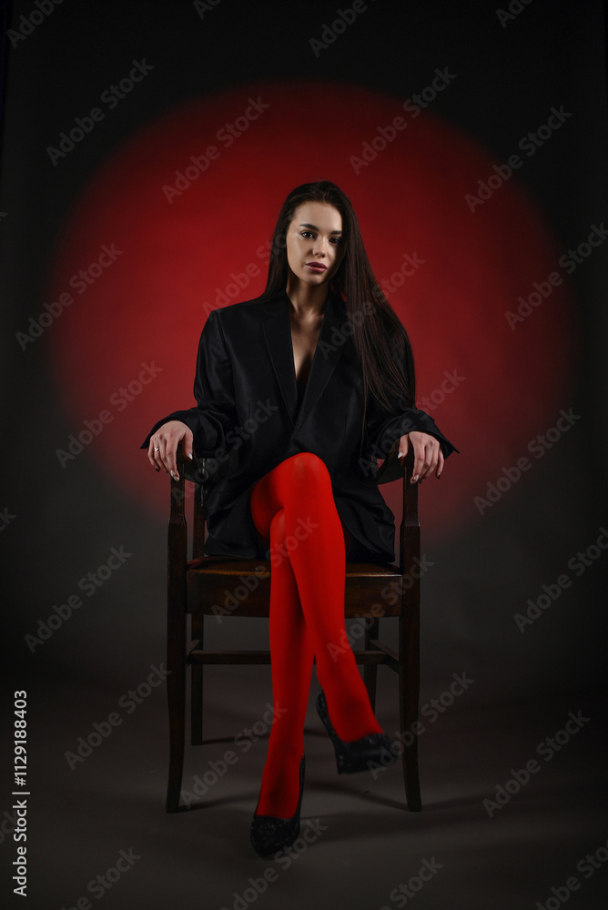 Beautiful girl in a black oversized blazer and red tights posing on the chair on a dark background. Beautiful girl in a black oversized blazer and red tights posing on the chair on a dark background.