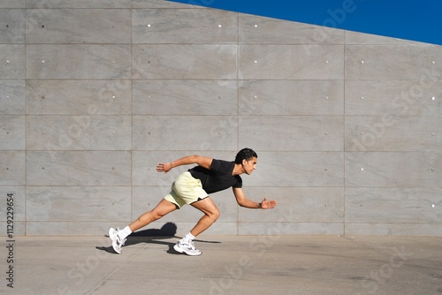 A man in a black shirt and yellow shorts leans forward into a running stance against a gray concrete wall outdoors under a bright blue sky photo