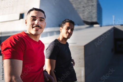 A man in a red shirt and a man in a black shirt smile while standing outdoors in a relaxed and sunny environment with modern architectural buildings in the background