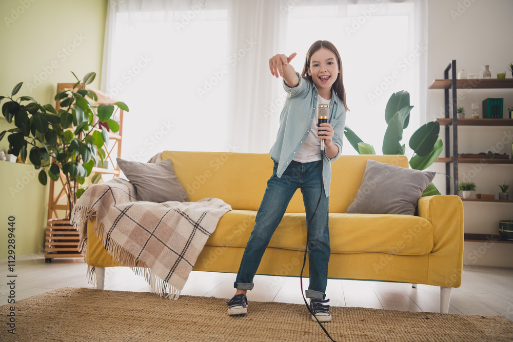 Obraz premium Joyful young girl singing with microphone in living room, wearing casual denim jeans and shirt, enjoying a leisure day indoors at home