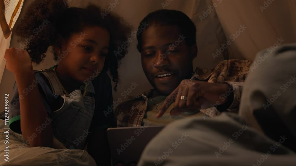 Medium close up shot of little biracial girl and his Black dad watching funny videos on tablet computer sitting inside teepee tent at home at night