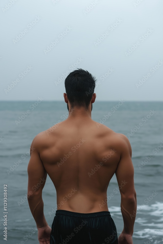 A man with a muscular back is standing on a beach with the ocean in the background. The man is in a relaxed and confident mood, enjoying the view of the ocean A man with a muscular back is standing on a beach with the ocean in the background. The man is in a relaxed and confident mood, enjoying the view of the ocean