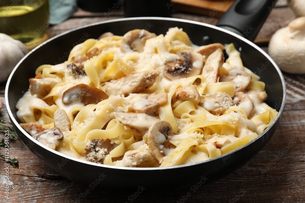 Tasty Alfredo pasta with chicken and mushrooms in frying pan on wooden table, closeup Tasty Alfredo pasta with chicken and mushrooms in frying pan on wooden table, closeup