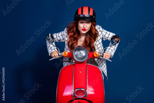 Dynamic young woman with red hair and lipstick rides a red scooter against a dark blue background in a trendy checkered suit