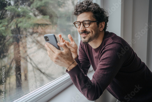 Young handsome middle eastern man with glasses having a video call on mobile phone while standing near window.