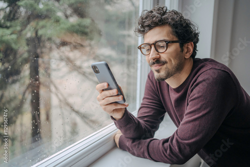 Fotografía Young handsome middle eastern man scrolling on mobile phone in the morning standing near window