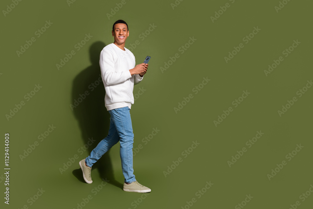 Young afro-american man smiling and walking forward holding smartphone against khaki background in white sweater and jeans Young afro-american man smiling and walking forward holding smartphone against khaki background in white sweater and jeans