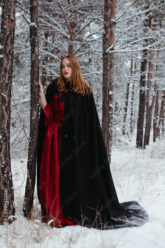 girl in red dress and black cloak walking in snowy forest. woman in medieval dress in winter forest. girl in red dress and black cloak walking in snowy forest. woman in medieval dress in winter forest.