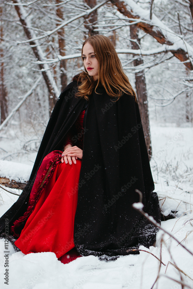 girl in red dress and black cloak walking in snowy forest. woman in medieval dress in winter forest. girl in red dress and black cloak walking in snowy forest. woman in medieval dress in winter forest.