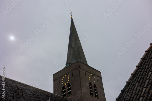 A tall church steeple with a clock and a weather vane stands against a cloudy gray sky, partially surrounded by rooflines, creating a serene and timeless architectural scene.