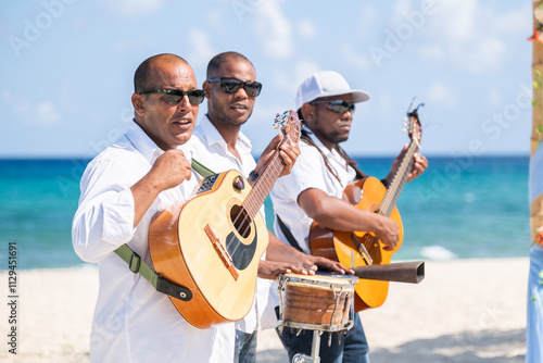 Three black musicians dressed in suits are standing on caribbean coastline and playing musical instruments guitar, drums, violin near arch at wedding ceremony	