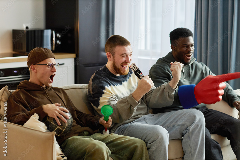 Group of three excited male friends watching game on TV at home and holding sports fan accessories Group of three excited male friends watching game on TV at home and holding sports fan accessories