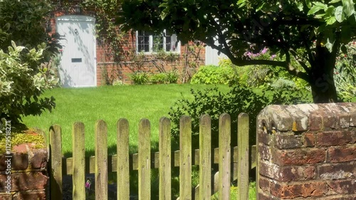 Charming cottage garden with a neatly maintained lawn, brick exterior, and a white door, framed by leafy trees and climbing vines in a quaint English village. High quality 4k footage