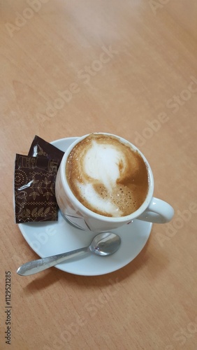 Uma xícara branca de capuccino com dois pacotinhos pretos de açúcar, pires branco e colher, sobre mesa marrom. photo