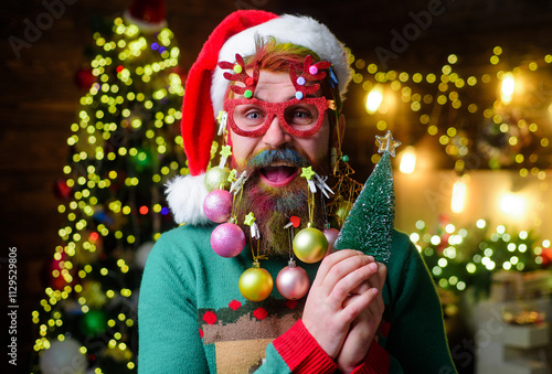 Merry Christmas. Happy bearded man in Santa hat and party glasses with little decorative Christmas tree. Handsome bearded man with painted hair and beard in party glasses holds small Christmas tree. photo