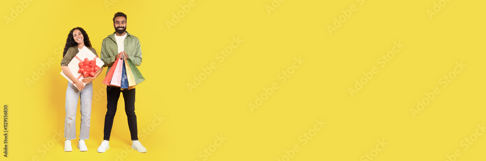 Gifts Shopping. Happy Middle Eastern Spouses Holding Shopper Bags And Wrapped Present Box Standing On Yellow Studio Background, Smiling At Camera. Holiday Sales Season Offer Concept, Copy Space Gifts Shopping. Happy Middle Eastern Spouses Holding Shopper Bags And Wrapped Present Box Standing On Yellow Studio Background, Smiling At Camera. Holiday Sales Season Offer Concept, Copy Space