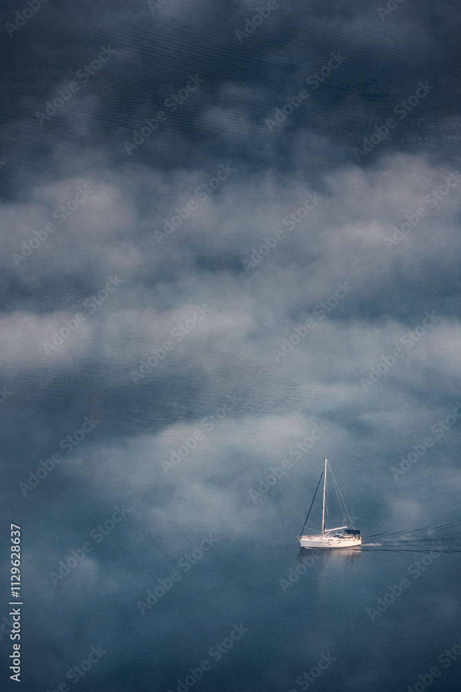 White sailboat sailing on calm sea with low clouds creating a mystical atmosphere