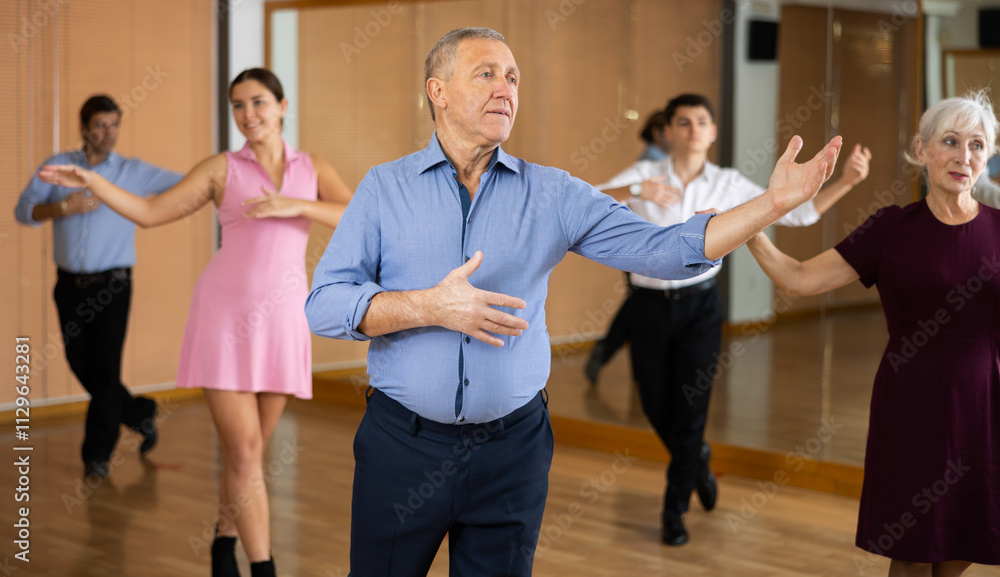 Fototapeta premium Positive elderly man practicing ballroom dance pose in training hall with other persons during dancing-classes
