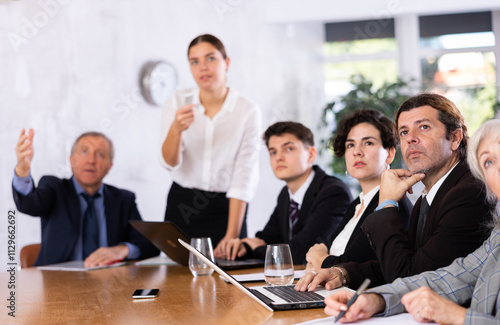 Office coworkers of different ages watching presentation while sitting at table with laptop and notes