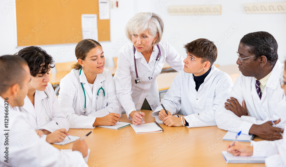 Group of medical workers in white coats sitting around desk during professional training course, listening to experienced aged female lecturer Group of medical workers in white coats sitting around desk during professional training course, listening to experienced aged female lecturer