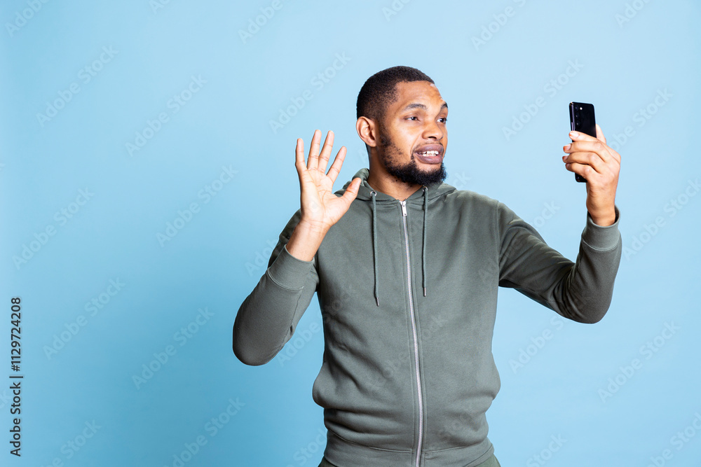 Young african american guy waving at video call conference in studio, chatting with his friends against blue background. Positive smiling person discussing on remote online connection. Young african american guy waving at video call conference in studio, chatting with his friends against blue background. Positive smiling person discussing on remote online connection.