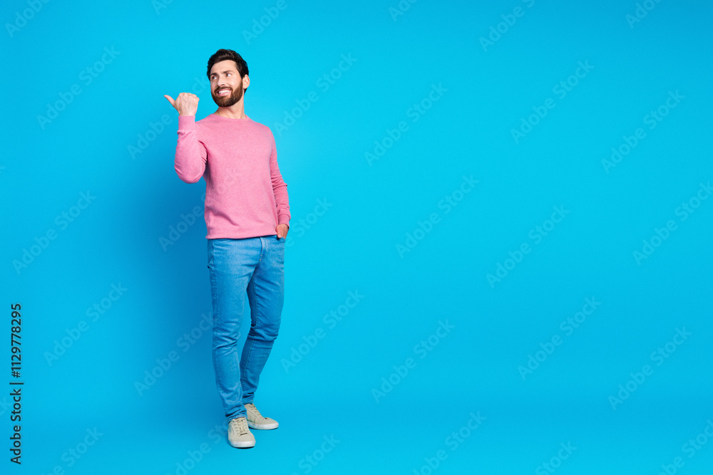 Confident man pointing to the side wearing casual pink sweater and blue jeans on a vibrant blue background Confident man pointing to the side wearing casual pink sweater and blue jeans on a vibrant blue background