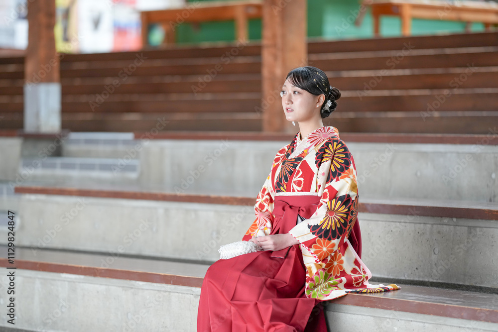 Japanese woman in her 20s wearing red hakama, staple for Japanese university graduates, sits in audience seats in large room. Japanese woman in her 20s wearing red hakama, staple for Japanese university graduates, sits in audience seats in large room.