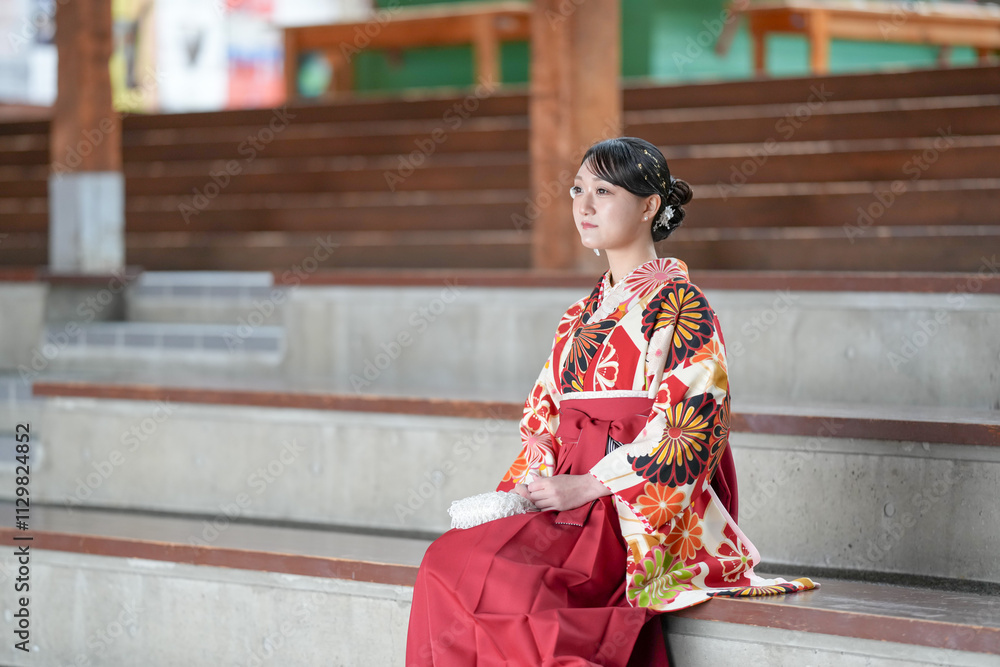 Japanese woman in her 20s wearing red hakama, staple for Japanese university graduates, sits in audience seats in large room. Japanese woman in her 20s wearing red hakama, staple for Japanese university graduates, sits in audience seats in large room.