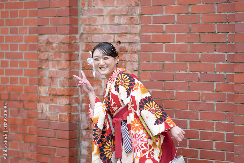 Fototapeta premium A Japanese woman in her 20s, wearing a red hakama, a staple of Japanese university graduates, poses against a cultural brick building.