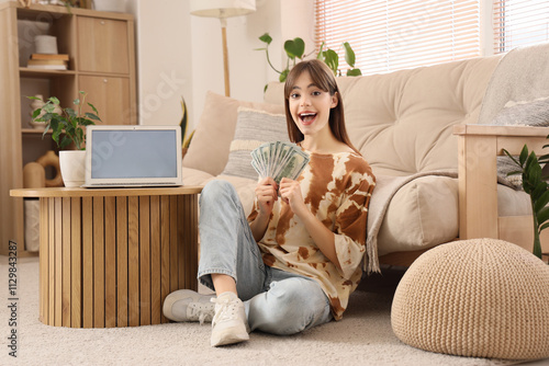 Young woman with dollar banknotes and blank laptop on table at home