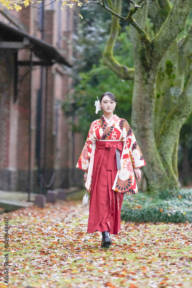 A Japanese woman in her 20s walks in front of a large tree and traditional Japanese buildings, wearing a red kimono (hakama), a staple for Japanese university graduates. A Japanese woman in her 20s walks in front of a large tree and traditional Japanese buildings, wearing a red kimono (hakama), a staple for Japanese university graduates.